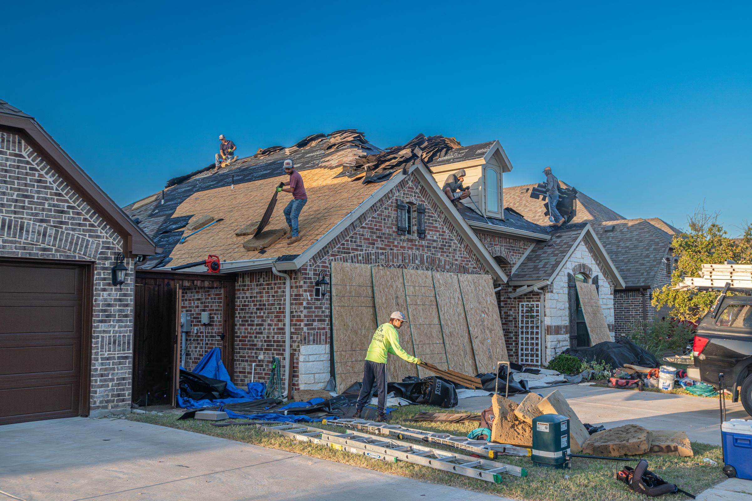 Kestrel crew tearing off a hail-damaged roof on a brick home; new plywood decking leans against the garage